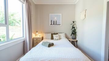 A neatly made bed with a rolled towel, a bedside table with a lamp, a framed picture on the wall, and a potted plant.