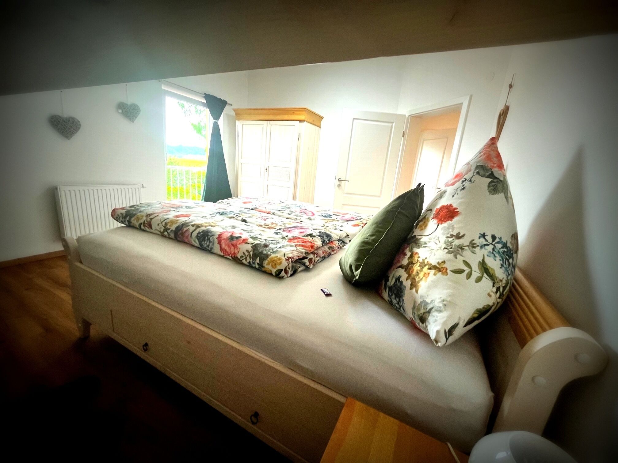 A single bed with a floral bedspread and two pillows, a radiator, a window with curtains, and a door.