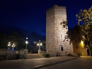 Ein historischer Steinturm mit einer Treppe, umgeben von einer gepflasterten Fläche mit Laternen und einem Gebäude mit steinverkleideter Fassade.