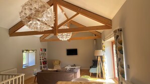 A spacious room with a high ceiling, wooden beams, a wooden dining table, white chairs, a sofa, a bookshelf, and a television. - Daisy Barn - Beautiful Grade 2 listed conversion (Mevagissey)