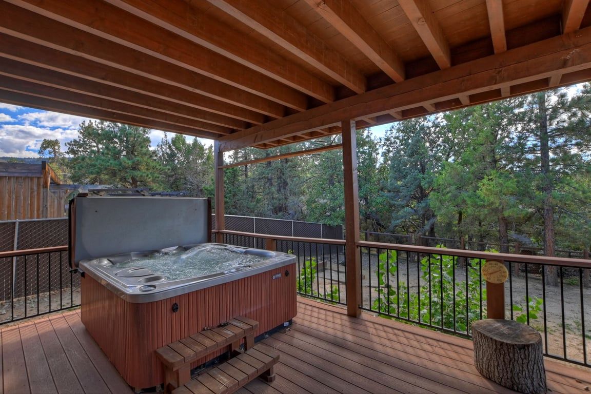 A wooden deck with a hot tub, wooden benches, and a railing overlooking a forested area.