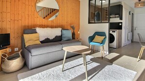 A modern living room with a grey sofa, a small round table, and a blue armchair. A mirror hangs on the wall, and there is a staircase leading to another level.