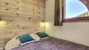 A room with wooden walls, a bed with patterned bedding, a window with curtains, and a small wall-mounted light.