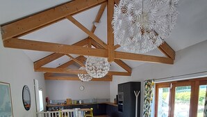 A spacious living area with a high ceiling, wooden beams, a white sofa, a round table, and a kitchen in the background. - Blossom Barn - Beautiful Grade 2 listed barn conversion (Mevagissey)