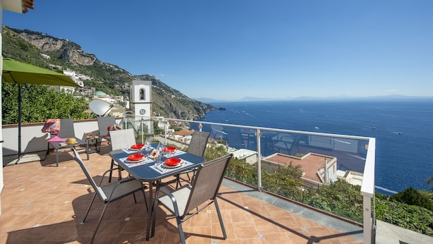 A terrace with a table set for dining, overlooking a coastal view with a church and mountains in the background.