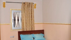 A bedroom with a wooden bed, blue pillows, a red bedspread, a small bedside table, a nightstand, a window with curtains, and a ceiling light fixture.