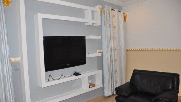A living room with a flat-screen TV mounted on a white wall, a black leather sofa, and a patterned curtain.