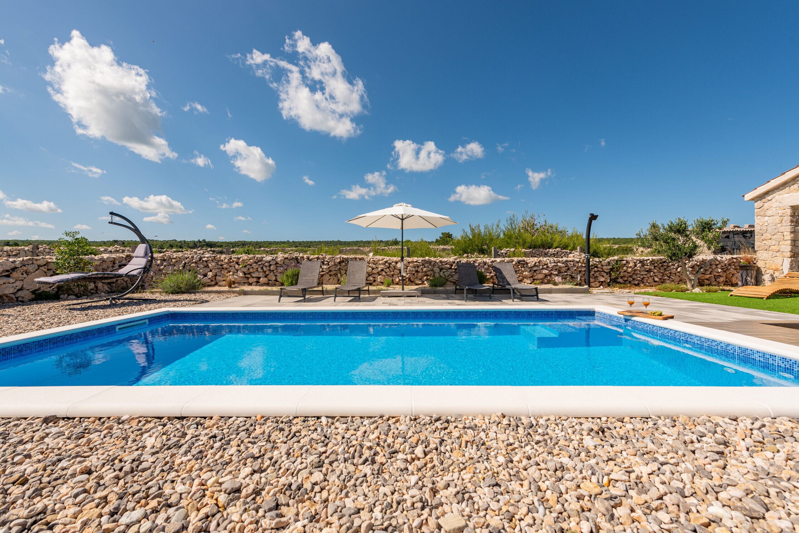 Piscina rectangular con borde de guijarros, rodeada de sillas de descanso y una sombrilla blanca.