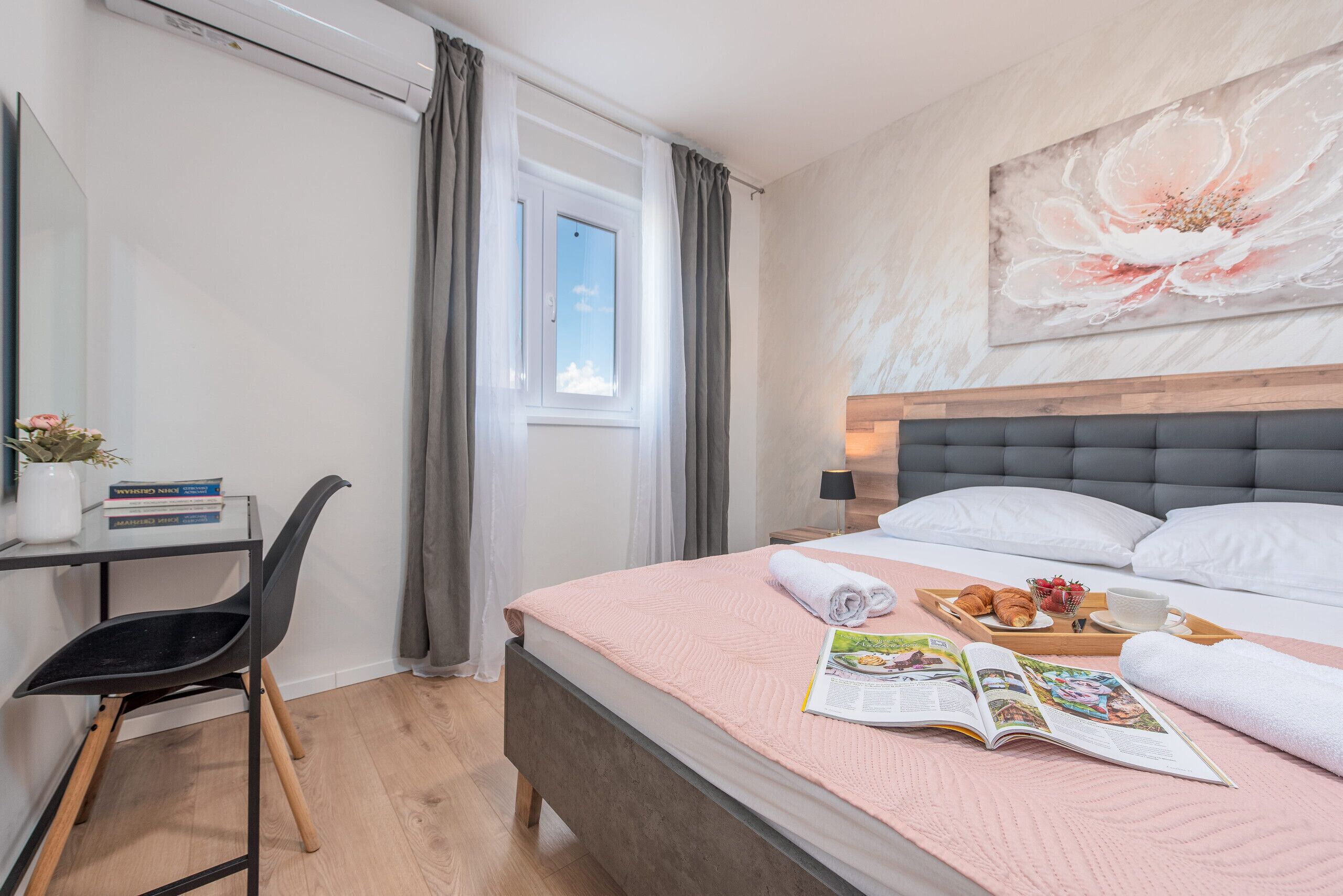 Une chambre bien rangée, avec un lit, une table de chevet, une chaise, une fenêtre avec des rideaux et un grand tableau floral accroché au mur.