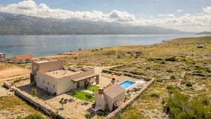 Una casa moderna con piscina, rodeada por un muro de piedra y vistas a un lago y montañas.