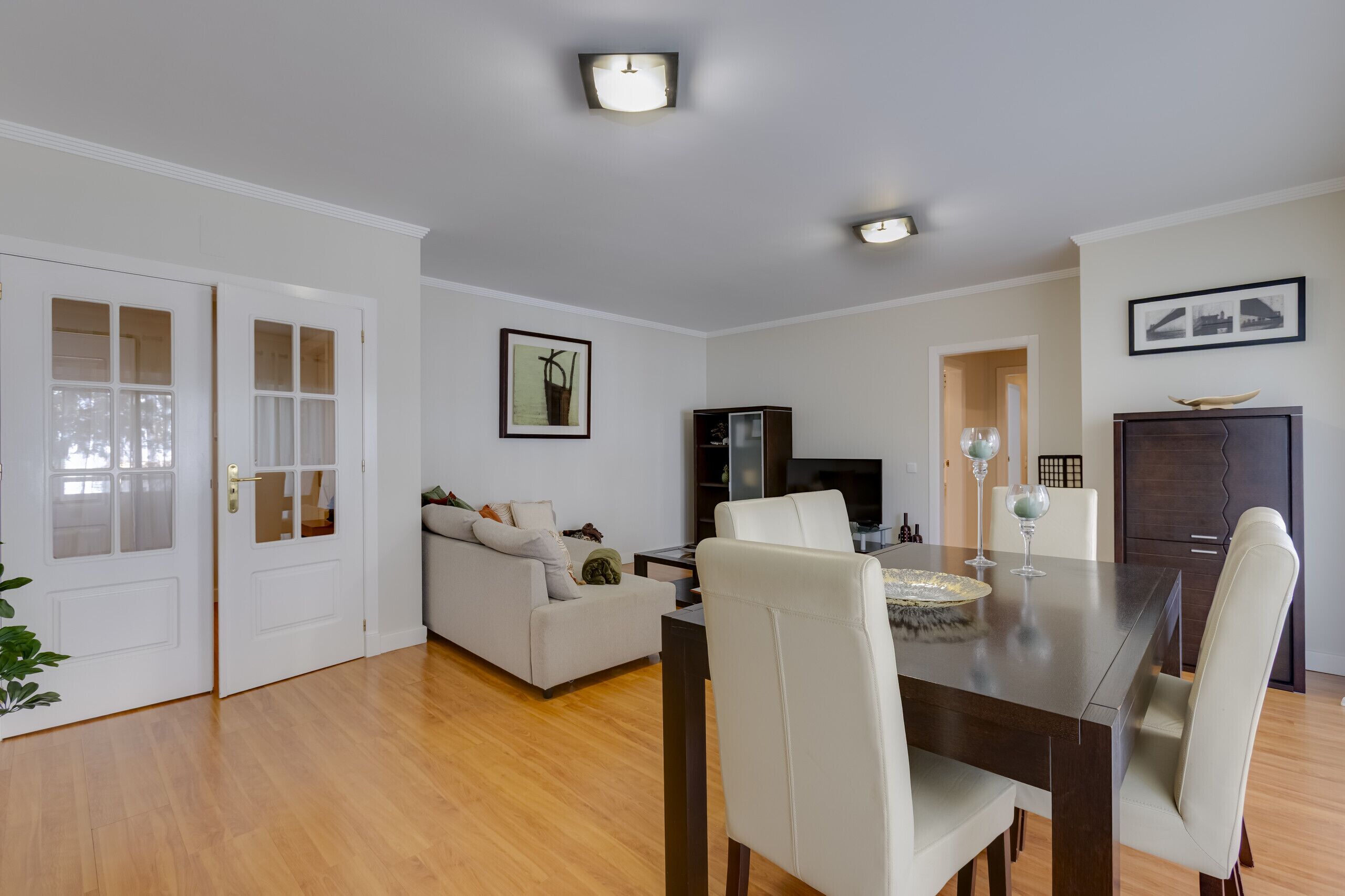 Een moderne woonkamer met een eethoek, met een houten vloer, witte muren en een donkerhouten eettafel met witte stoelen.