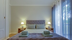 A neatly made bed with a headboard, two bedside tables with lamps, and a view of the outdoors through a window with curtains. - Jazz & Silvia (Albufeira)