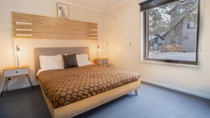 Une chambre à coucher avec un grand lit, deux tables de chevet, une tête de lit dotée d’un éclairage intégré, une fenêtre donnant sur un paysage enneigé et un tableau encadré accroché au mur.