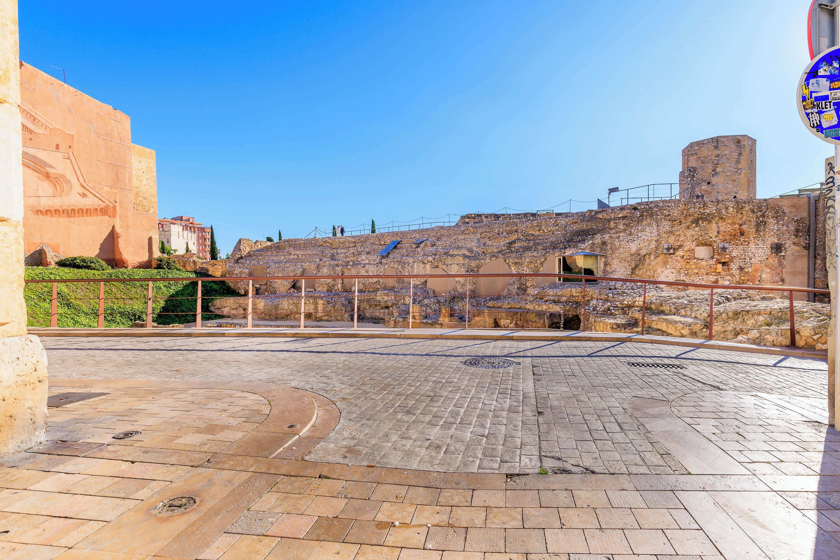 A paved courtyard with a stone wall, a metal railing, and a building with a tower in the background.