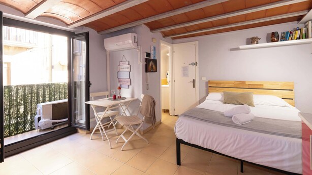 A bedroom with a bed, a table, and a chair, a balcony with a view, and a door leading to a bathroom.