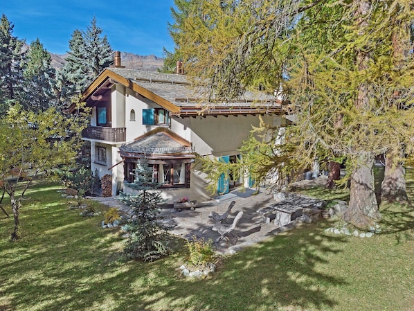 Ein zweigeschossiges Haus mit einer Steinterrasse, einem kleinen Gazebo und einem großen Baum im Vordergrund.