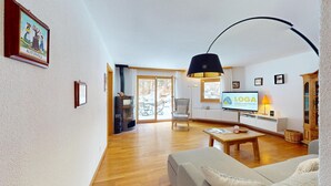 A spacious living room with a wooden floor, a sofa, a TV, and a fireplace. - Beautiful 2-room apartment on the raised ground floor with approx. (Champfèr)