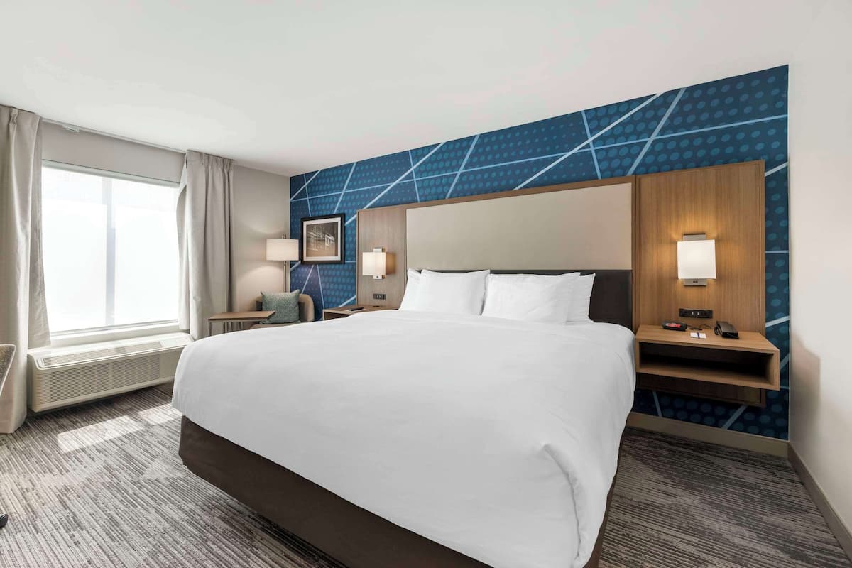 A hotel room with a large bed, a desk, a chair, a nightstand, and a wall-mounted lamp.