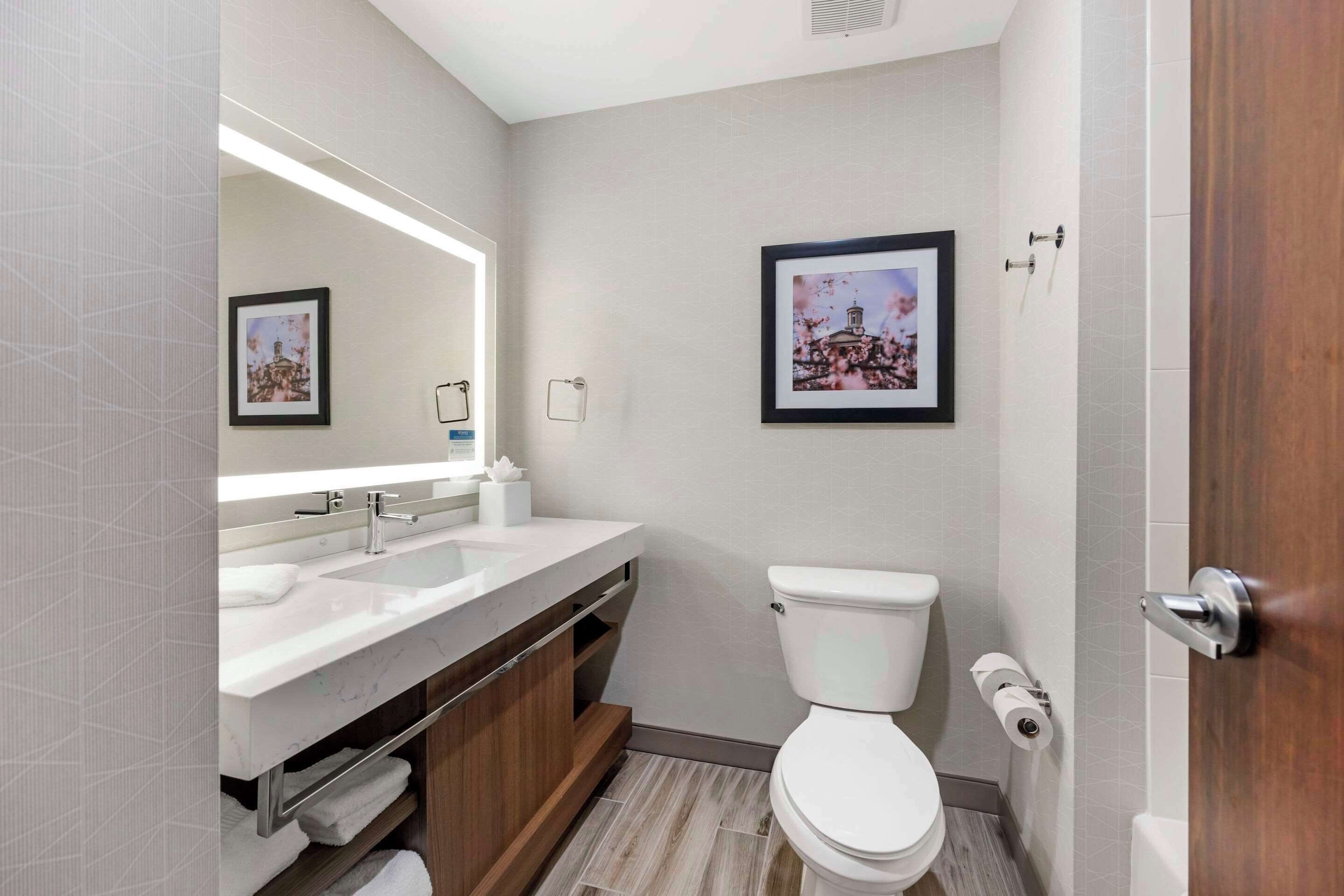 Een moderne badkamer met een wit wastafelmeubel, spiegel en toilet. Twee ingekaderde foto's hangen aan de muur, een handdoekhouder en een houten kastje.