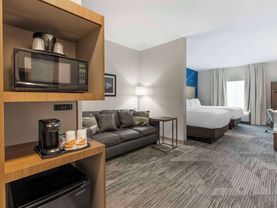 A hotel room with a microwave, coffee maker, and storage shelves. It includes a sofa, a bed, a desk, and a lamp.
