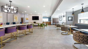 Een moderne hotellobby met een lange receptiebalie, paarse barstoelen, groene stoelen en een zithoek met gestreepte stoelen en een bank.