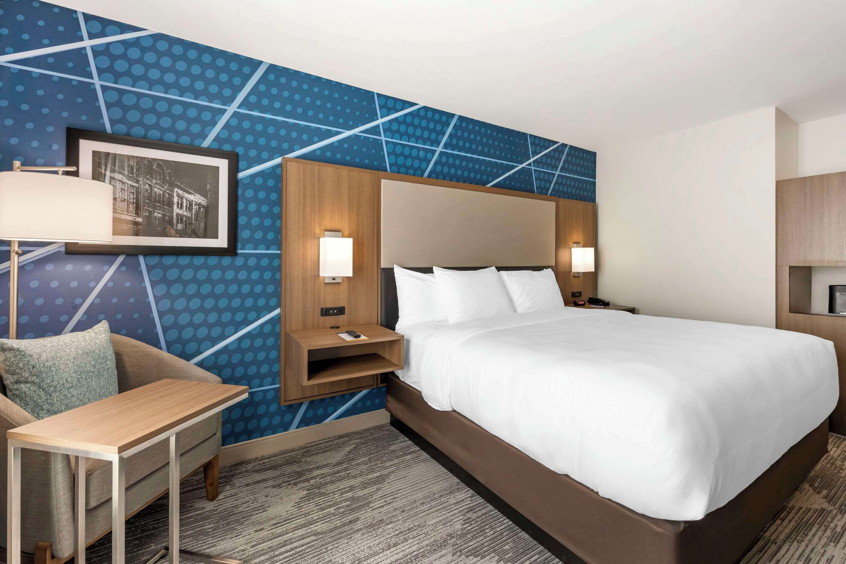 A hotel room with a large bed, a desk, and a chair. There is a wall-mounted picture and two bedside lamps.