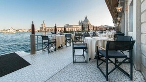 Un área de comedor al aire libre con mesas y sillas, con vista a un cuerpo de agua y edificios históricos.