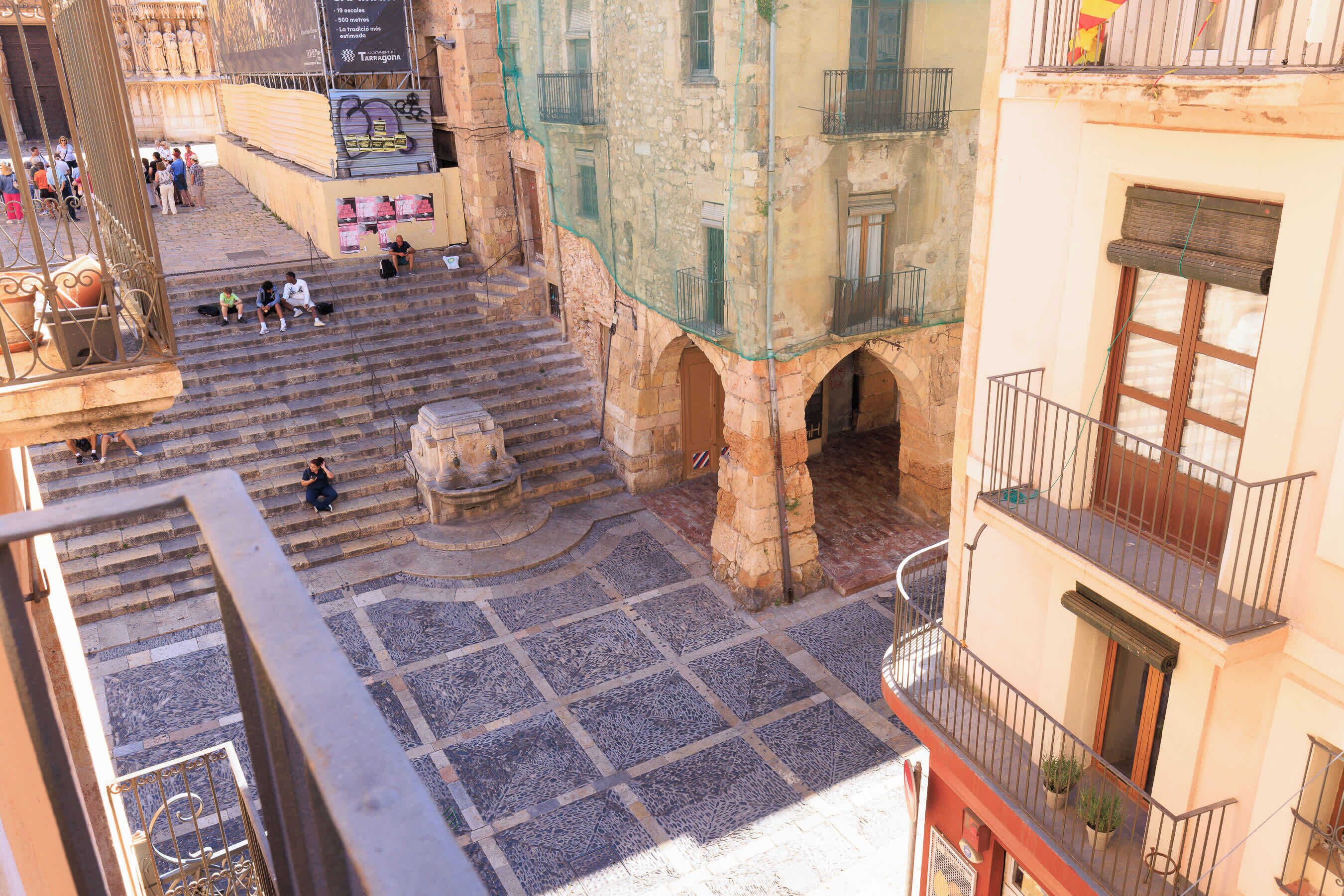 A courtyard with stone steps, a fountain, and buildings with balconies.