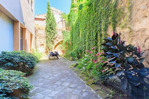 A narrow stone pathway leading to a seating area with a table and chairs, surrounded by lush greenery and a stone wall.