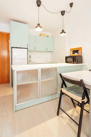 A compact kitchen with a refrigerator, a microwave, a dining table with a chair, and two pendant lights.