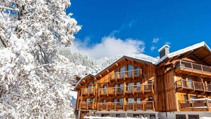A wooden chalet with multiple balconies, surrounded by snow-covered trees and a clear blue sky.