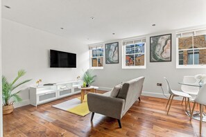 A modern living room with a flat-screen TV, a grey sofa, a white coffee table, and a glass dining table with chairs.