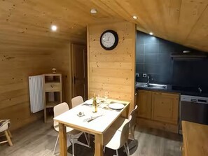 Ein Holzinterieur mit einem Esstisch für zwei Personen, einer Wanduhr und einer Küchenzeile mit modernen Geräten.