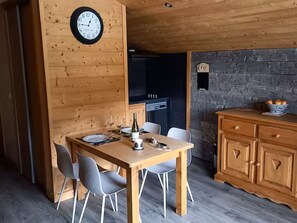A wooden dining area with a clock, a table set for two, and a stone wall in the background.