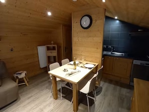A wooden interior with a dining table set for two, a clock on the wall, and a kitchen area.