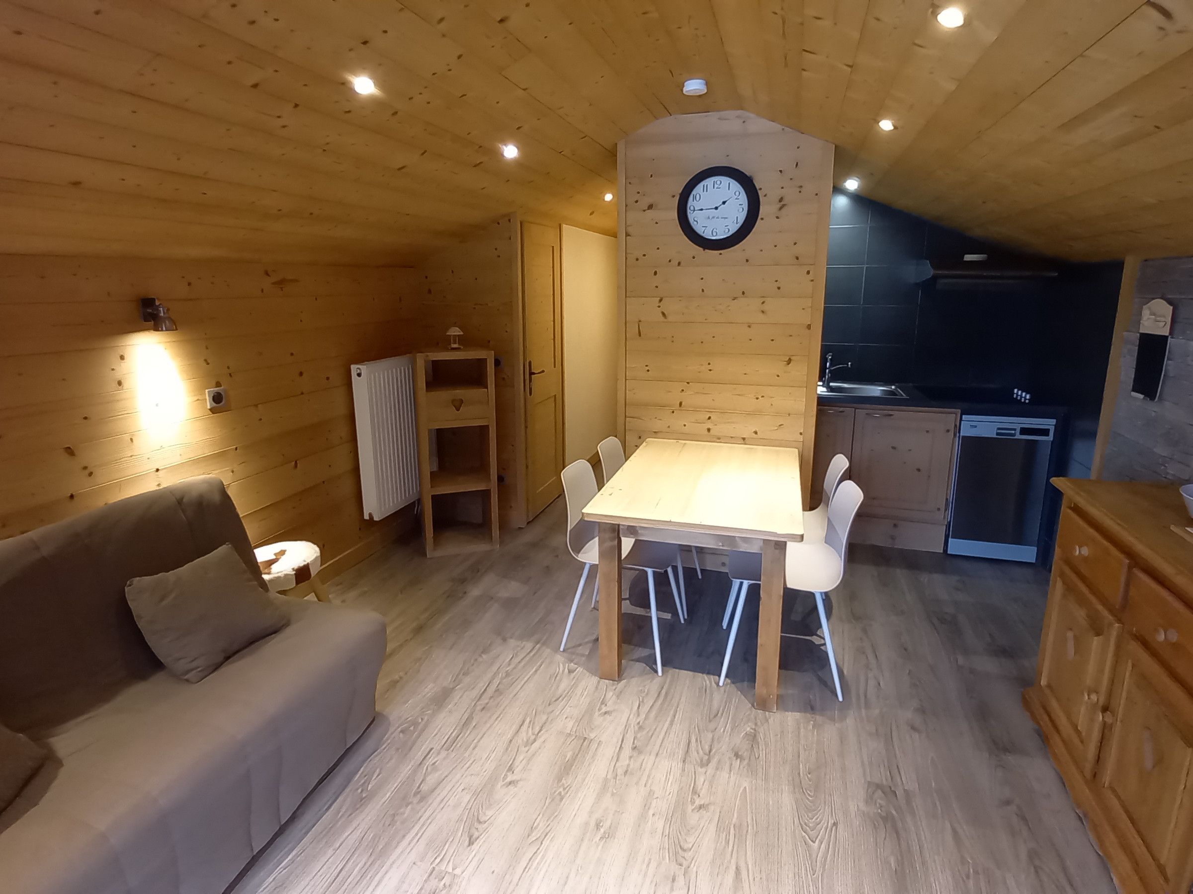 A cozy cabin interior with a wooden dining table, a sofa, a clock on the wall, and a kitchen area.