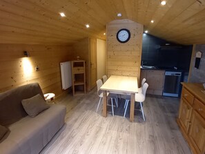 A cozy cabin interior with a wooden dining table, a sofa, a clock on the wall, and a kitchen area.