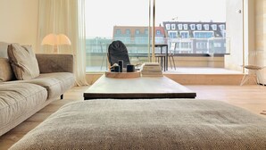 A modern living room with a sofa, a coffee table with books, a chair, and a view of buildings through large windows. - Beautiful Specious Design Duplex Penthouse With sea View (Knokke-Heist)
