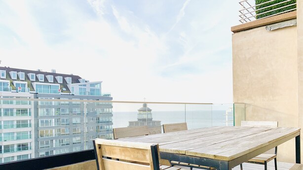 A balcony with a wooden table and chairs, overlooking a cityscape and the sea. - Beautiful Specious Design Duplex Penthouse With sea View (Knokke-Heist)