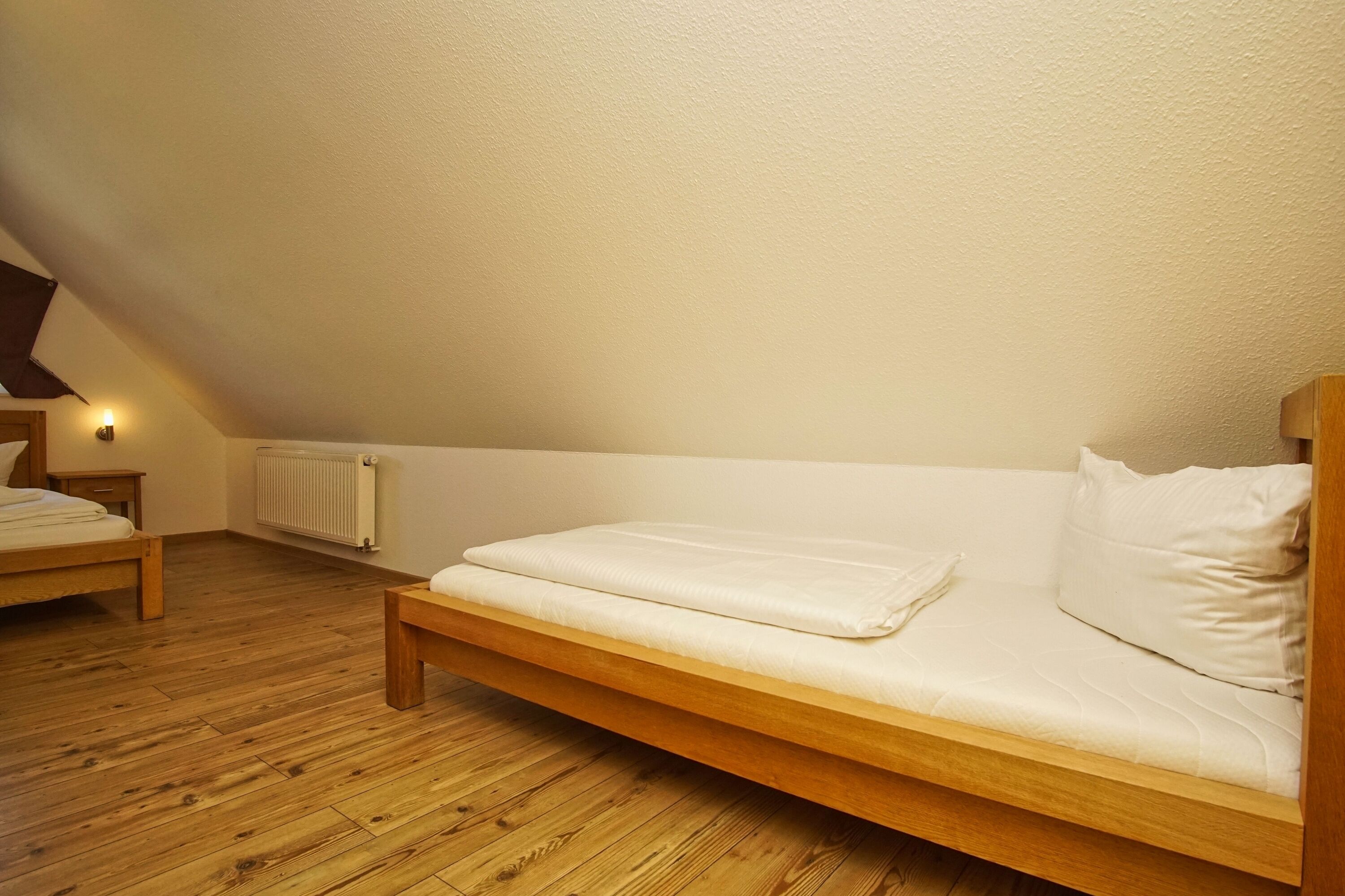 Una cama individual con estructura de madera y ropa de cama blanca en una habitación con suelo de madera y techo inclinado.