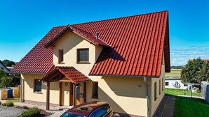 Une maison à deux étages, avec un toit recouvert de tuiles rouges, une véranda en bois et une voiture garée dans l’allée.