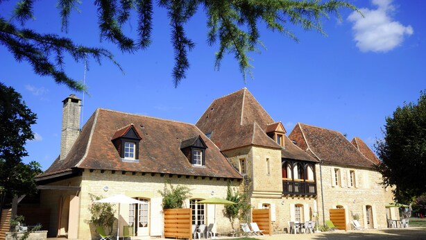 A traditional stone building with multiple chimneys, a covered patio with outdoor seating, and a well-maintained lawn. - Le Clos des Rives Appt N°07 (Saint-Vincent-de-Cosse)