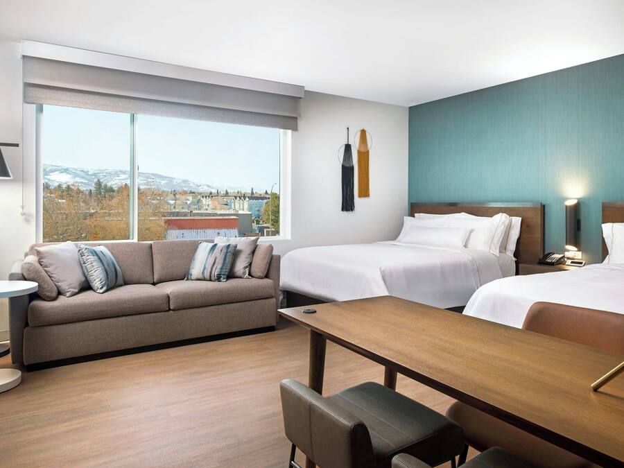 A hotel room with a large window, a sofa, two beds, a wooden table, and a desk.