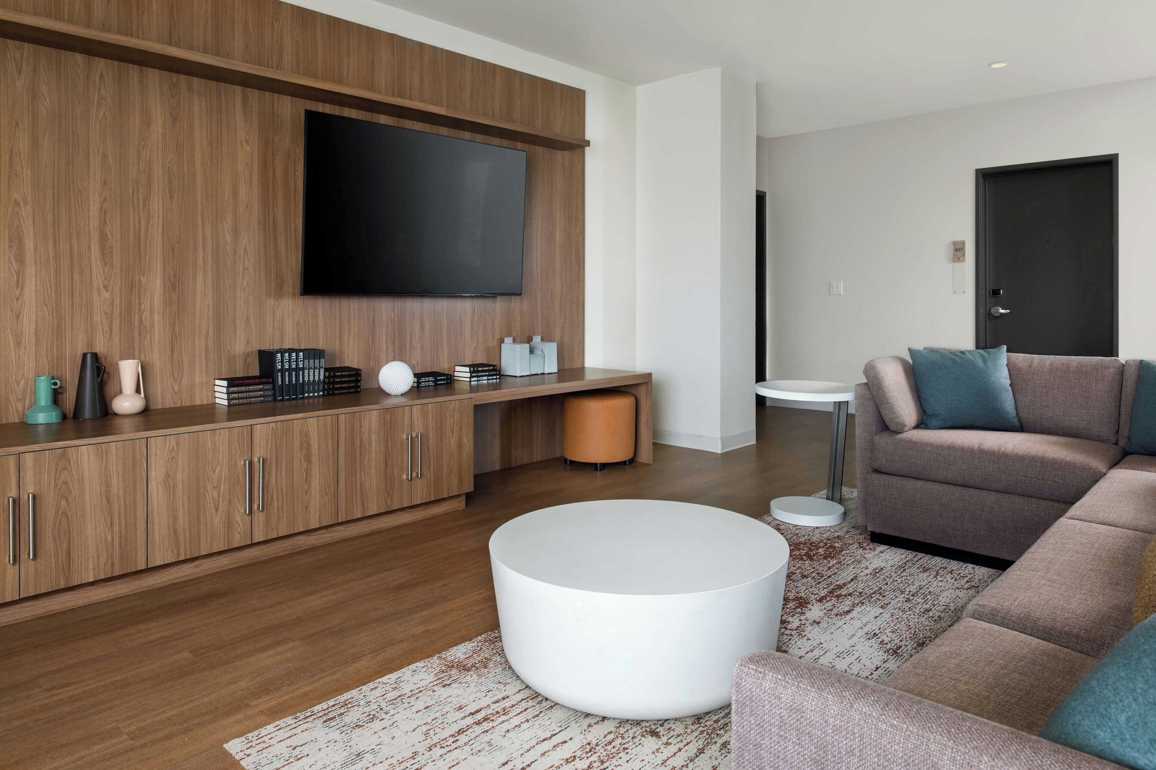 Een moderne woonkamer met een houten kast, een flatscreen televisie, een ronde witte salontafel en een hoekbank.