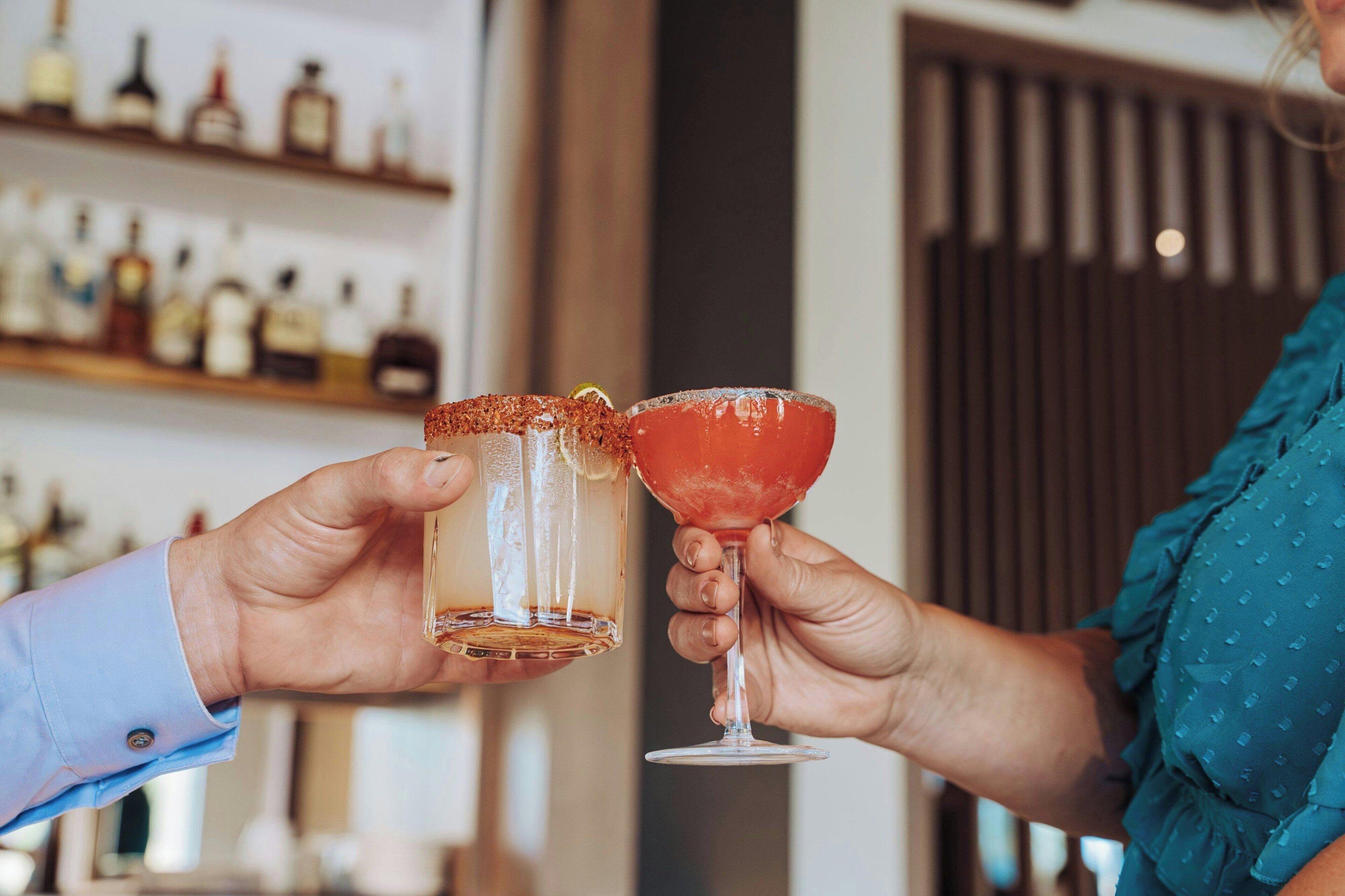 Two hands holding cocktail glasses, one with a clear liquid and the other with a red liquid, in a bar setting with shelves of bottles in the background.