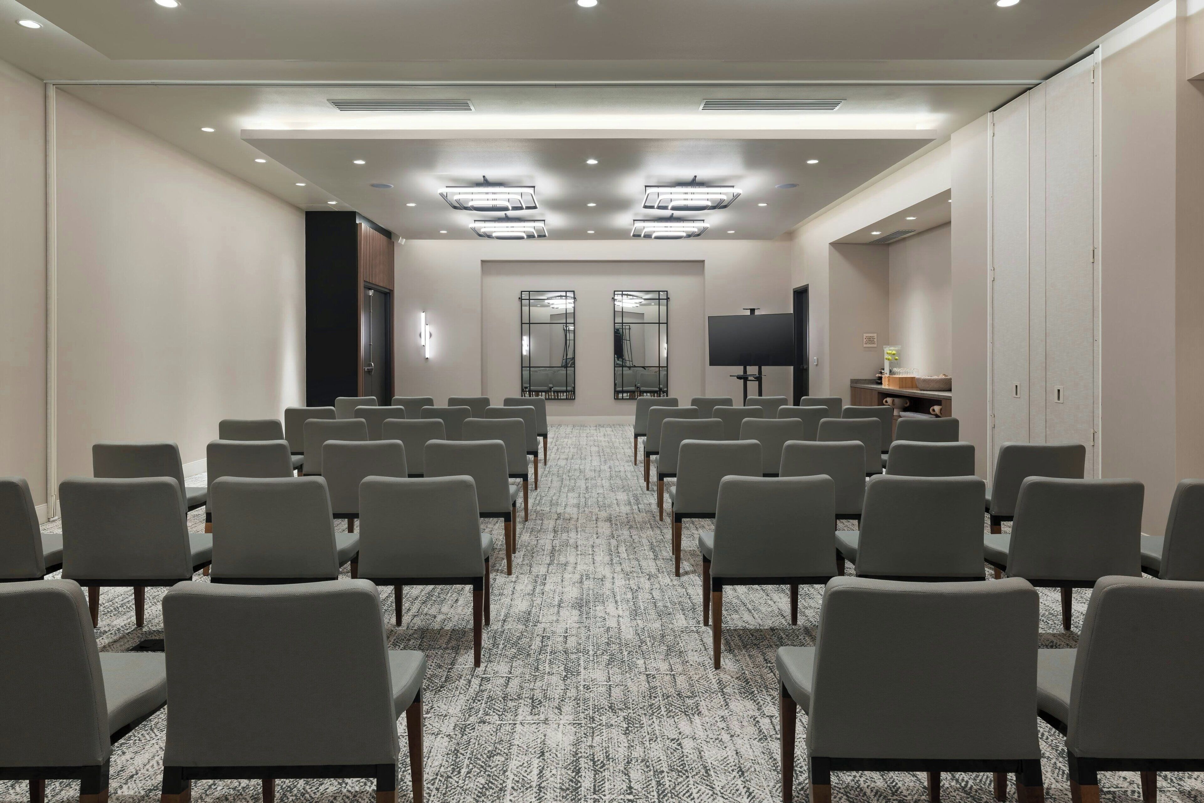 A conference room with rows of chairs, a carpeted floor, a flat-screen TV, and a door at the far end.