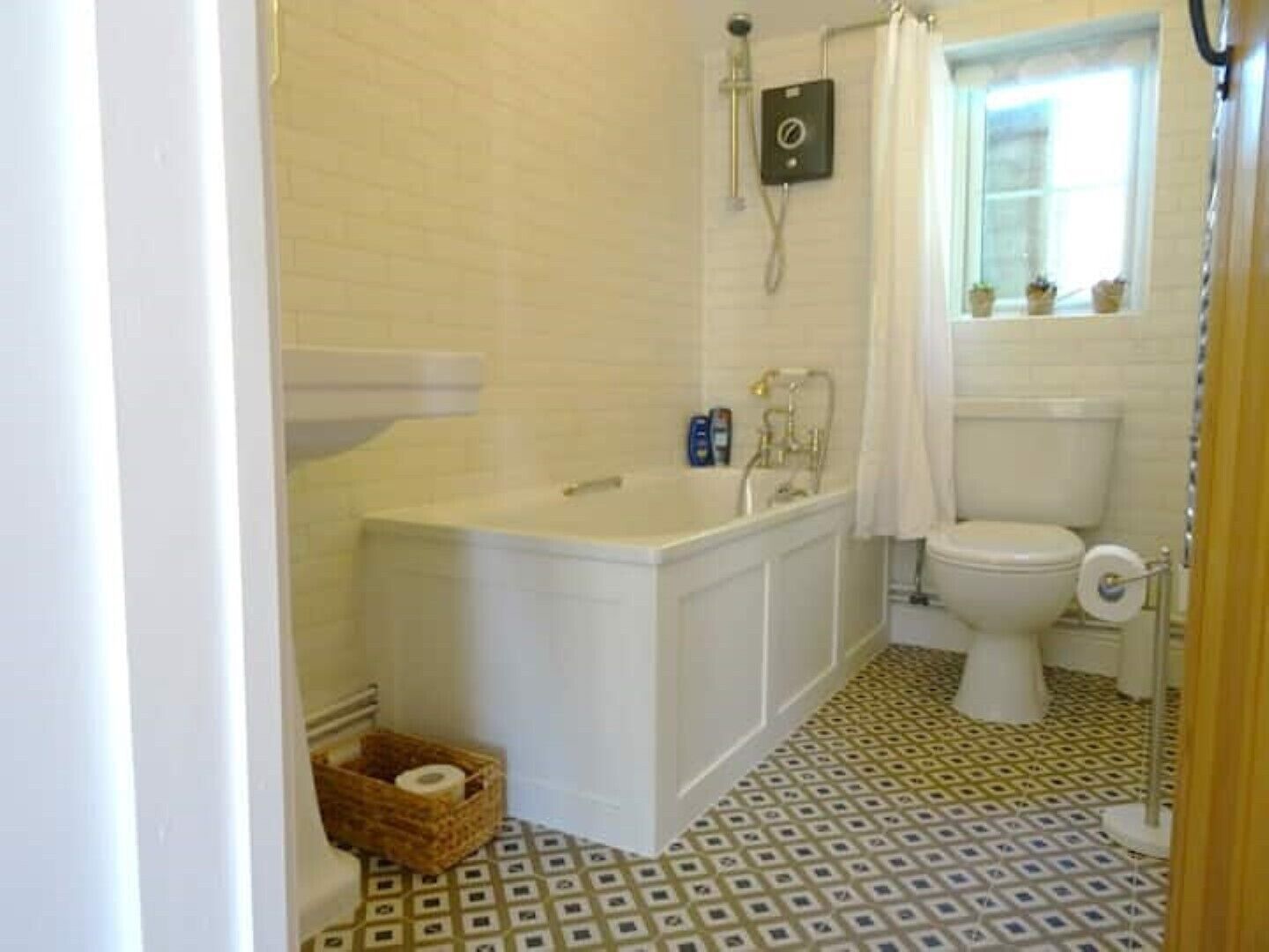 Une salle de bain avec une baignoire blanche, des toilettes et une douche. Le sol est carrelé et il y a un panier contenant un rouleau de papier toilette.