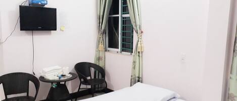 A small room with a bed, two chairs, a small table, a television mounted on the wall, and a window with curtains.