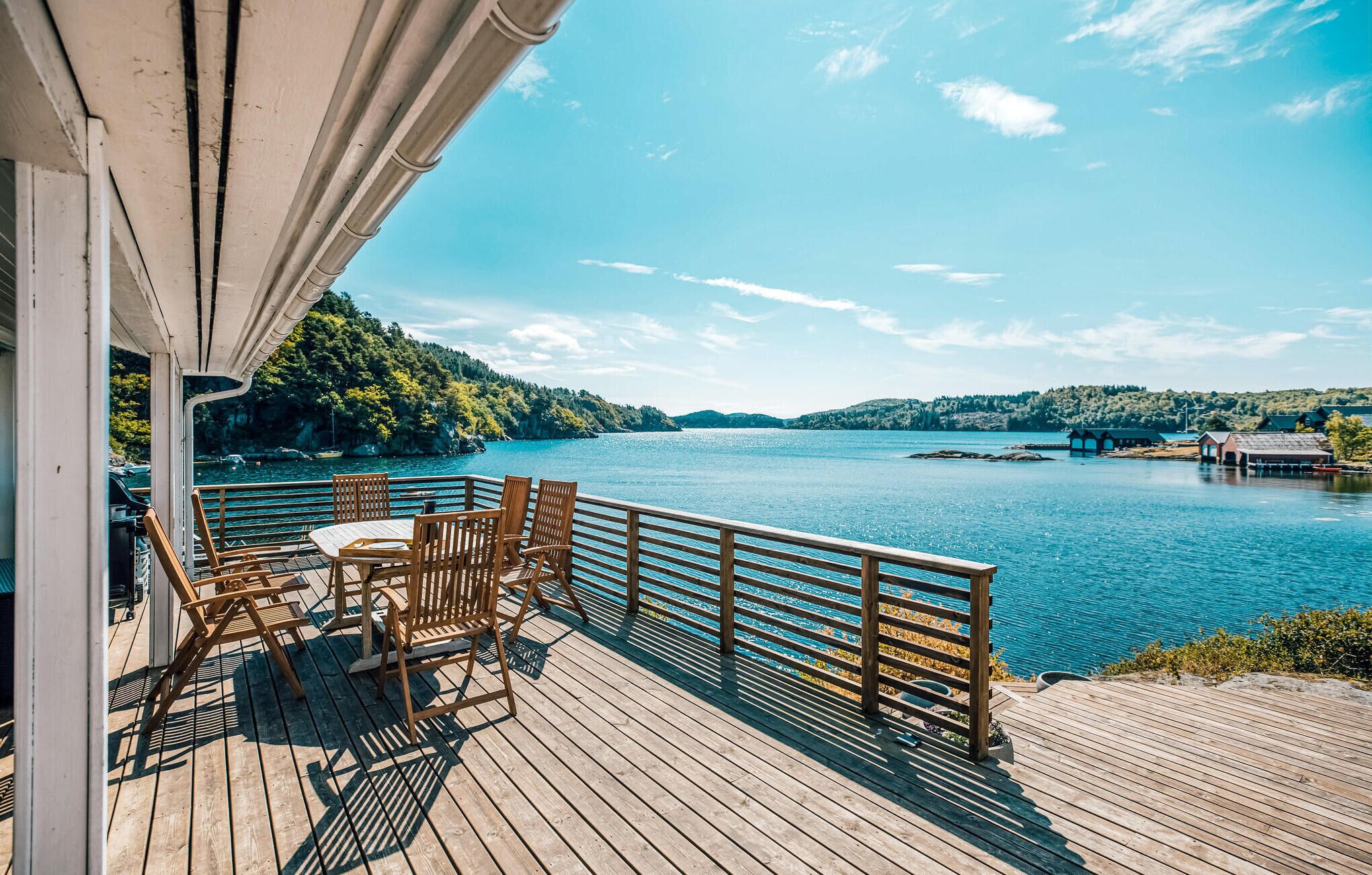 Une terrasse en bois avec une table et des chaises, d’où l’on a une vue sur un lac paisible, avec des bateaux et des bâtiments visibles au loin.
