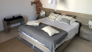 Une chambre à coucher comprenant un lit, des tables de chevet, une commode, une œuvre d’art accrochée au mur et une plante suspendue.